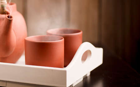 Two clay colored ceramic mugs, hot with steam on a white wooden tray and a teapot or coffee pot with a rustic wooden backgroundの写真素材