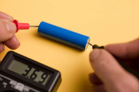 battery 18650 with volt calculator, voltmeter in the hands of a man, red positive pole, black negative pole, on a yellow work tableの写真素材