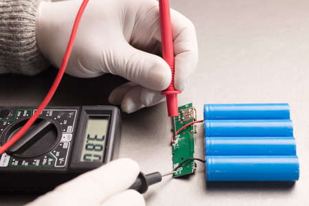 Charge indicator circuit, multimeter red and black poles in the hands of man checking voltage and amps, 18650 blue batteries, gray background. powerbank.の写真素材
