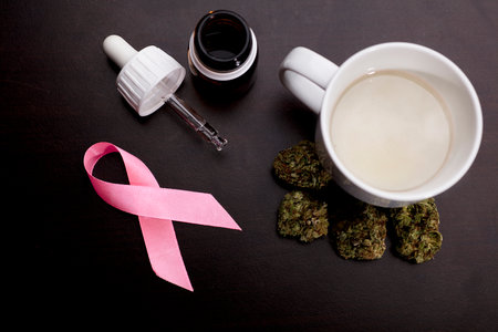 cancer pink ribbon with marijuana buds, white cup with infusion steam, oil drops dispenser bottle, on brown table, wooden backgroundの写真素材