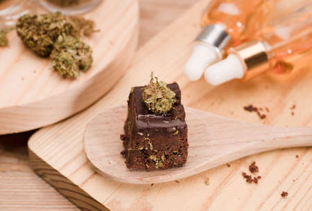 Chocolate cupcake muffins with cannabis buds in wooden spoon, oil pot and cooking jars. Marijuana hemp in food dessert. Chocolate herb muffins cooking. Marijuana cake on a brown wooden table.の写真素材