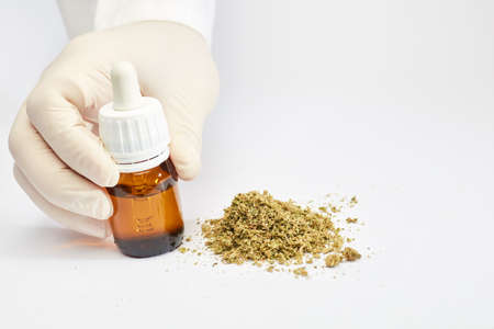 marijuana oil dropper bottle in hands of doctor with crushed marijuana in pile, white backgroundの写真素材