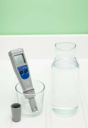 pH meter in a glass of water, and bottle on a white and green background. Measurement of the characteristics of drinking water. The hardness of the water. Drinking water quality analyzer.の写真素材