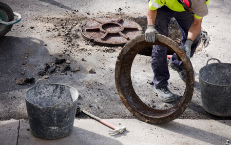 sewage and manhole in hands of worker in uniform and asphalt at construction siteの写真素材