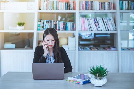 Young asian employee is very busy with her tasks. She is recieving call while she using laptop at...