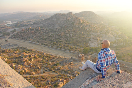 A handsome bald man in glasses sits on a sunset or dawn background in Hampi, on the top of the mountain.の写真素材