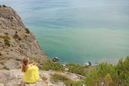 A young girl in a yellow bright dress sits on top of a mountain, against a turquoise and blue world. Loneliness and travel in nature. Crimea.の写真素材