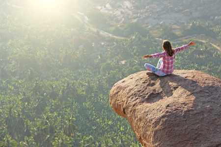 A young girl in a lotus pose is greeting the sun on a background of sunrise or sunset sitting on top of a mountain or on a large rock.の写真素材