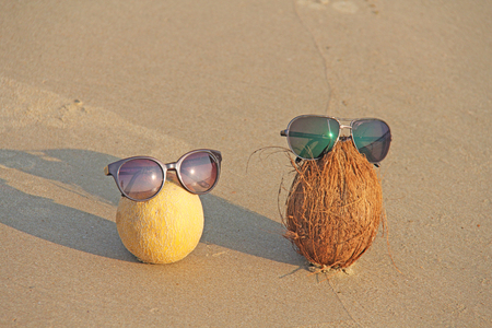 Orange cut melon with seeds inside and a brown coconut with sunglasses, on the beach and on the sea, against the background of sand. Asian tropical fruits. Fruit art and humor. Design with copy space.の写真素材