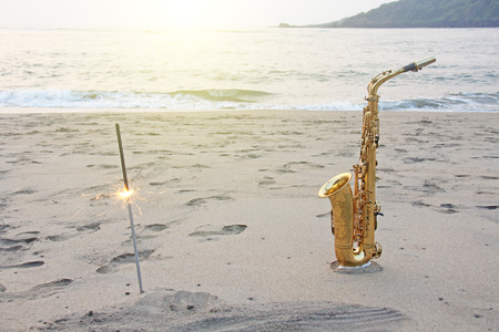 The golden saxophone alto stands on the sand and on the shore, against the background of the sea and Bengal lights. Romantic musical background. Musical cover and holiday. Design with copy space.の写真素材