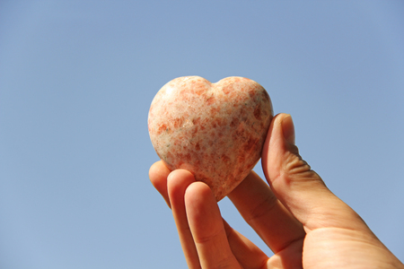 A hand holds a heart made of natural stone rhodochrosite against a blue sky. Heart in hand. A stone in the shape of a heart. Love talisman, zen, spa.の写真素材