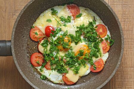 Fresh fried eggs are cooked in a frying pan, with tomatoes, cheese and greens. Vegetarian dishes. Bright healthy food on a wooden background, rustic.の写真素材