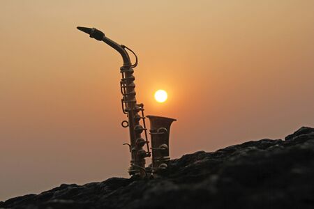 The golden saxophone alto stands on a black stone, against a background of sunset. Silhouette of saxophone against the sky. Romantic musical. Musical cover and creative. Design with copy space.の写真素材