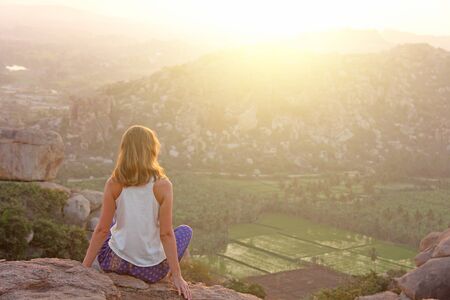 A girl sits with her back on the top of the mountain and looks at the sunset at Hampi and welcomes the sun. Meditation, harmony, alone with nature, silence. Large stones and rice fields in Hampi.の写真素材