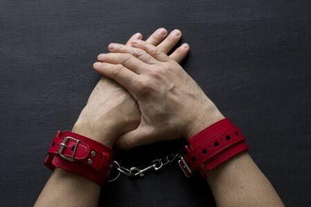 Adult male hands in red erotic leather handcuffs. Hands in captivity. Red leather handcuffs on black...
