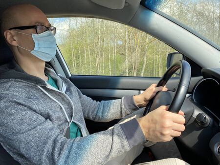 Man in medical protective mask driving car, close-up. Hygienic mask to prevent infection, airborne respiratory illness such as flu, Covid - 2019. Protection against coronavirus, contagious disease.の写真素材