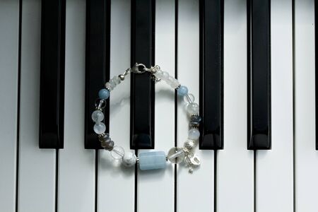 A handmade blue bracelet made of natural stones, aquamarine, cacholong, labrador, moonstone, with heart and key pendants, lies on black and white piano keys. Asymmetric arm bracelet.の写真素材