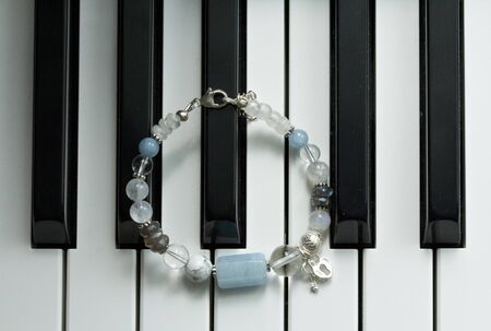 A handmade blue bracelet made of natural stones, aquamarine, cacholong, labrador, moonstone, with heart and key pendants, lies on black and white piano keys. Asymmetric arm bracelet.の写真素材