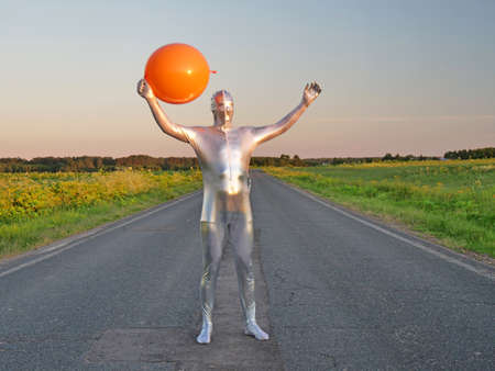 Tall slender man in silver suit is fully dressed in tight silver suit, stands in summer on an empty road, track and holds large bright orange balloon. Freak, an alien, wonderful unusual person, UFO.の写真素材