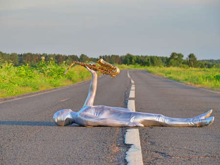 Man musician saxophonist in full body-hugging silver and silver electric suit holding golden alto saxophone, lies on empty road in summer with perspective. Freak, unusual person, alien, UFO.の写真素材
