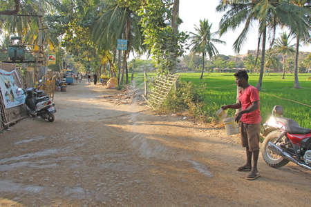 India, Hampi, 02 February 2018. The main street of the village of Virupapur Gaddi. Guesthouses overlooking the rice fields. An Indian man sprinkles water on the road. Drops of water in flight.のeditorial素材