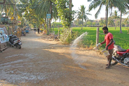 India, Hampi, 02 February 2018. The main street of the village of Virupapur Gaddi. Guesthouses overlooking the rice fields. An Indian man sprinkles water on the road. Drops of water in flight.のeditorial素材