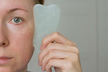 Facial massage. A young girl holds a white jade gouacha scraper for a natural mineral and makes a facial massage, close-up. Therapeutic healing massage for beauty and youth. Copy space for your text.の写真素材