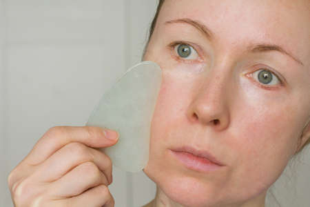 Facial massage. A young girl holds a white jade gouacha scraper for a natural mineral and makes a facial massage, close-up. Therapeutic healing massage for beauty and youth. Copy space for your text.の写真素材