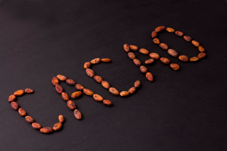 Word cocoa is laid out on black background made from natural unrefined cocoa beans. Raw materials for making cocoa powder, cocoa beverages, chocolate. Health drink concept. Flat lay, copy space.の写真素材