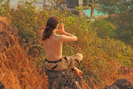 India, GOA, January 23, 2018. A man sits on top of a mountain and looks on the sea and on the sunset. A man in solitude, alone with nature, relax in nature. A man with his hands namaste.のeditorial素材