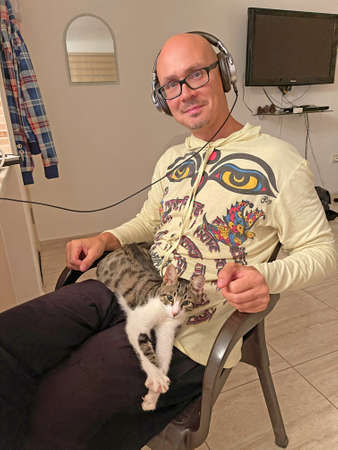 A beautiful cute kitten lies in the lap of a handsome male musician in headphones. True friendship, love, tenderness, affection and care between humans and animals.の写真素材