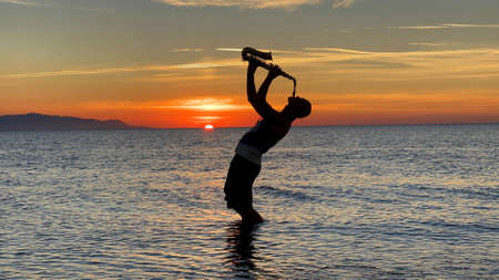Young male saxophonist stands with his feet in sea water, holds saxophone in his hands, looks at sunset. Beautiful sunset on sea, sky. Musician, playing saxophone, dancing, having fun, Silhouette.の写真素材