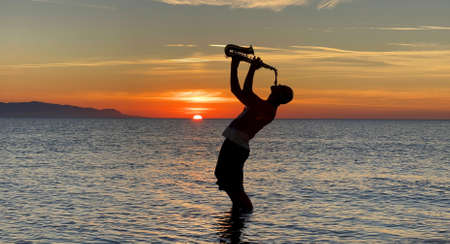 Young male saxophonist stands with his feet in sea water, holds saxophone in his hands, looks at sunset. Beautiful sunset on sea, sky. Musician, playing saxophone, dancing, having fun, Silhouette.の写真素材