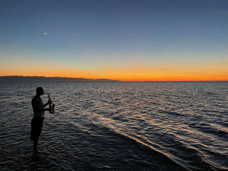 Young male saxophonist stands with his feet in sea water, holds saxophone in his hands, looks at sunset. Beautiful sunset on sea, sky. Musician, playing saxophone, dancing, having fun, Silhouette.の写真素材