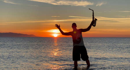 Young male saxophonist stands with his feet in sea water, holds saxophone in his hands, looks at sunset. Beautiful sunset on sea, sky. Musician, playing saxophone, dancing, having fun, Silhouette.の写真素材