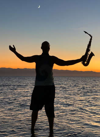 Young male saxophonist stands with his feet in sea water, holds saxophone in his hands, looks at sunset. Beautiful sunset on sea, sky. Musician, playing saxophone, dancing, having fun, Silhouette.の写真素材