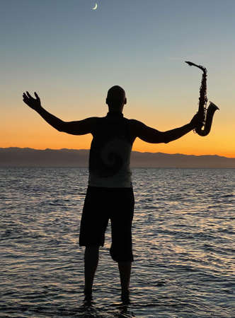 Young male saxophonist stands with his feet in sea water, holds saxophone in his hands, looks at sunset. Beautiful sunset on sea, sky. Musician, playing saxophone, dancing, having fun, Silhouette.の写真素材