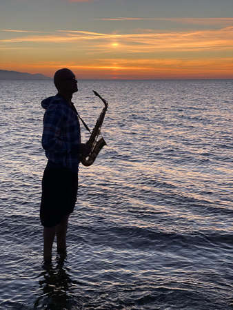 Young male saxophonist stands with his feet in sea water, holds saxophone in his hands, looks at sunset. Beautiful sunset on sea, sky. Musician, playing saxophone, dancing, having fun, Silhouette.の写真素材
