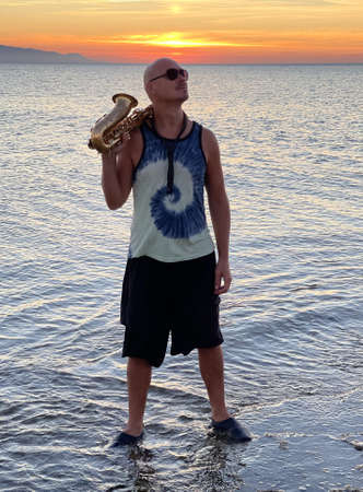 Young male saxophonist stands with his feet in sea water, holds saxophone in his hands. Beautiful sunset on sea, sky. Musician, playing saxophone, dancing, Silhouette. Wearing dark black glasses.の写真素材