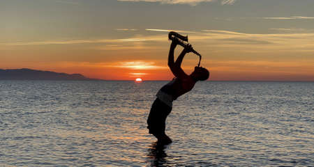 Young male saxophonist stands with his feet in sea water, holds saxophone in his hands, looks at sunset. Beautiful sunset on sea, sky. Musician, playing saxophone, dancing, having fun, Silhouette.の写真素材