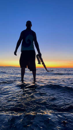 Young male saxophonist stands with his feet in sea water, holds saxophone in his hands, looks at sunset. Beautiful sunset on sea, sky. Musician, playing saxophone, dancing, having fun, Silhouette.の写真素材
