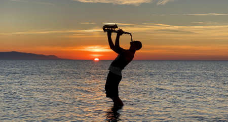 Young male saxophonist stands with his feet in sea water, holds saxophone in his hands, looks at sunset. Beautiful sunset on sea, sky. Musician, playing saxophone, dancing, having fun, Silhouette.の写真素材