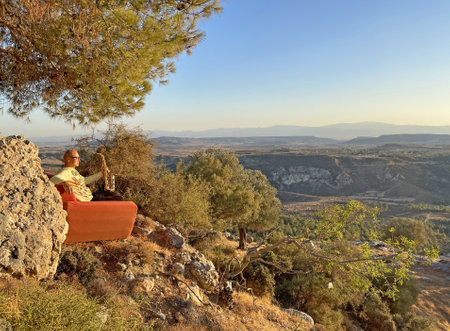 Young stylish bald male saxophonist sits on an orange sofa in mountains of Cyprus in nature, under pine tree, holds an alto saxophone in his hands. Musician with saxophone on nature in mountains.の写真素材
