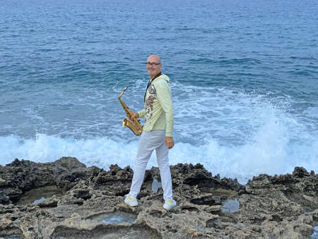 A young stylish bald male saxophonist stands against the backdrop of the sea and stormy high waves and plays the golden saxophone in Cyprus. Romantic Musician saxophonist.の写真素材