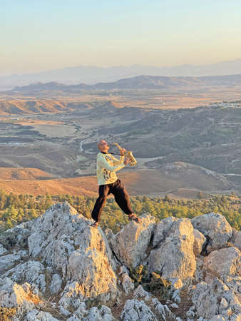 Young male saxophonist in black pants and yellow jacket, stands at the top of mountain against background of valley and holds saxophone musical instrument and plays. Height and beautiful nature.の写真素材