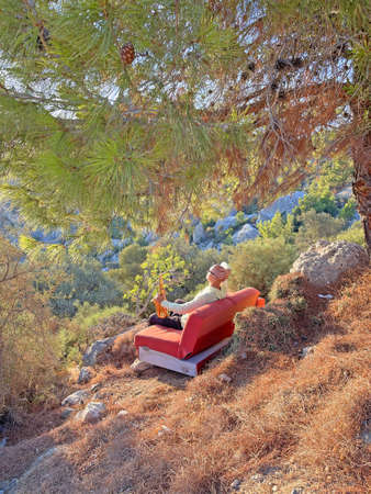 Young stylish bald male saxophonist sits on an orange sofa in mountains of Cyprus in nature, under pine tree, holds an alto saxophone in his hands. Musician with saxophone on nature in mountains.の写真素材