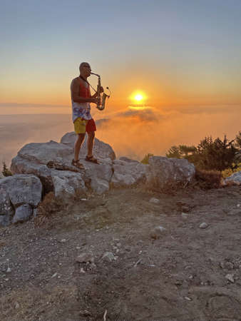 Young stylish bald male saxophonist stands on top of mountain against the backdrop of the sunset and the sun and plays the golden saxophone in Cyprus. Romantic Musician saxophonist. Above clouds.の写真素材