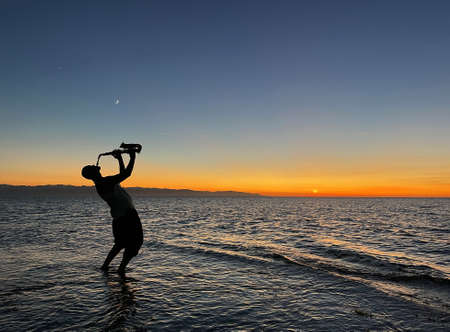 Young male saxophonist stands with his feet in sea water, holds saxophone in his hands, looks at sunset. Beautiful sunset on sea, sky. Musician, playing saxophone, dancing, having fun, Silhouette.の写真素材