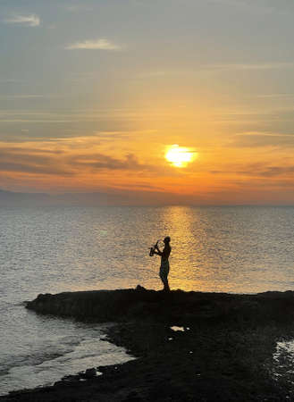 Young male saxophonist stands against background of sea and sunset, holds saxophone in his hands. Beautiful sunset on sea, sky. Musician saxophonist plays against background sea, sunset, silhouette.の写真素材