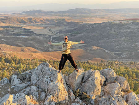 Young male saxophonist in black pants and yellow jacket, stands at the top of mountain against background of valley and holds saxophone musical instrument and plays. Height and beautiful nature.の写真素材